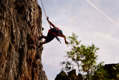 Low angle full length of active senior woman climbing on rocky cliff against sky