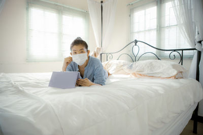 Full length portrait of woman wearing mask using laptop on bed