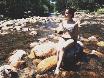 Full length of sensuous woman wearing bikini on rock amidst river at forest