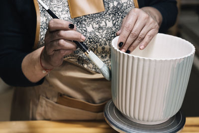 Crop anonymous female potter in apron painting handmade clay pot in professional workshop on blurred background