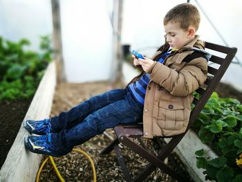 Side view of boy playing video game while sitting on chair in yard