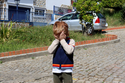 A red-haired boy stands alone in the middle of the street and covers his face with his hands.