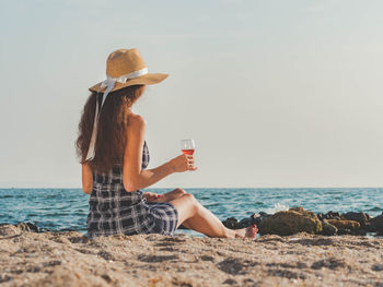 Woman having wine while sitting at beach against sky