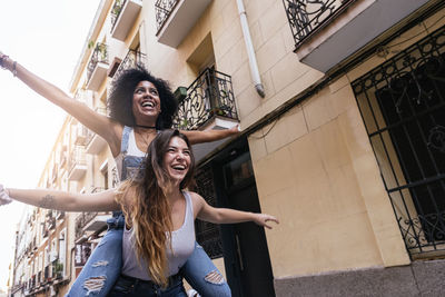 Smiling young woman with arms raised against building