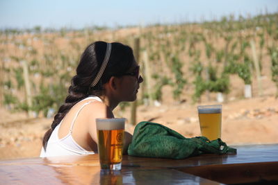 Side view of young woman by beer on table against vineyard