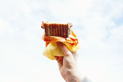 Cropped hand holding sandwich against cloudy sky