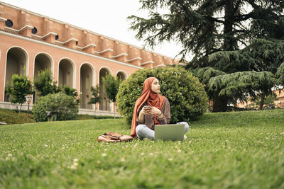 Muslim woman siting on the grass using a computer and phone