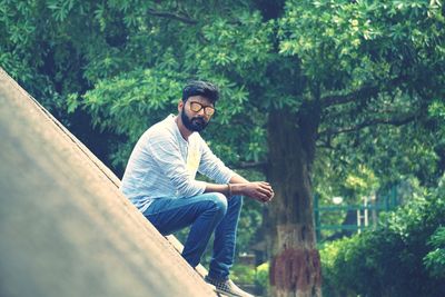 Young man wearing sunglasses sitting against trees in park