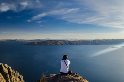 Rear view of woman sitting on cliff at sea against sky