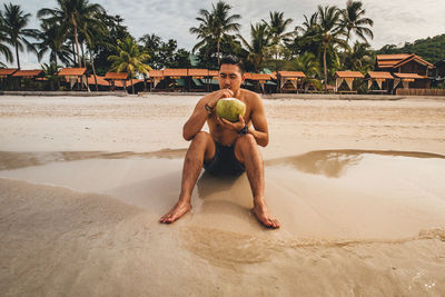 Full length of shirtless man drinking coconut water while sitting on shore at beach