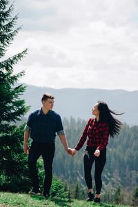 Full length of young couple holding hands while walking on field against sky during sunny day