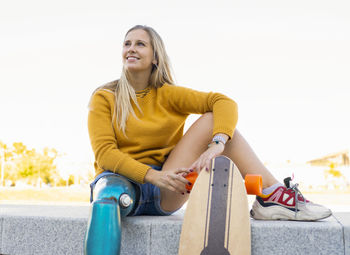 Carefree female skater in summer clothes and with bionic leg prosthesis sitting on stone border in city and looking away