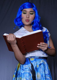 Young woman reading book against blue wall