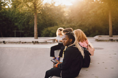 Thoughtful teenage girl holding mobile phone while sitting with friends at skateboard park