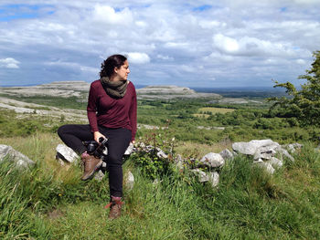 Young woman holding camera and sitting on rock at grassy field against cloudy sky