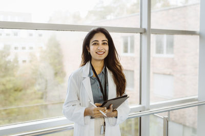 Portrait of smiling female doctor holding digital tablet and pen standing against window at hospital corridor