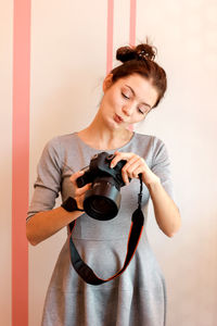 Young woman holding camera while standing against wall