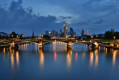 Illuminated bridge over river by buildings against sky at dusk