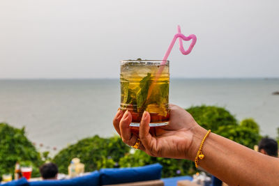 Cropped image of hand holding drink at sea against sky