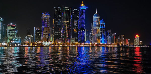 Illuminated buildings by river against sky at night