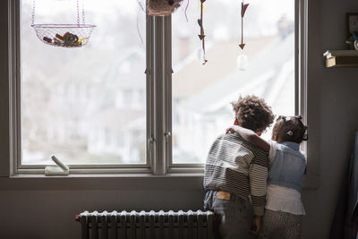 Rear view of siblings looking through window at home