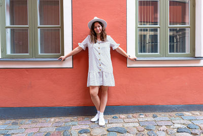 A young smiling woman in a white dotted dress with a stylish hat on the red wall of the building