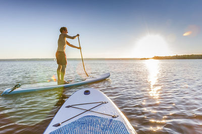 Side view of shirtless man paddleboarding on river against sky during sunset