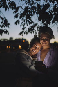 Smiling non-binary person using mobile phone by female friend at night
