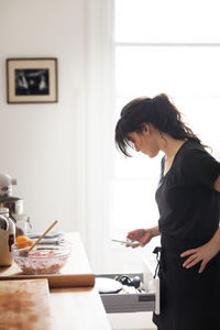 Side view of woman using smart phone while working in kitchen at home