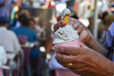 Man hand scoops gelato icecream from cup with background of outdoor street cafe diners.