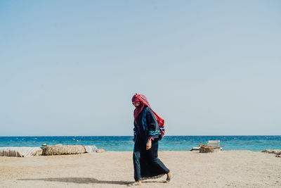 Side view of woman wearing traditional clothing while walking at beach against clear sky