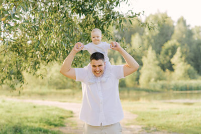 A young happy father of a large size holds his little son around his neck in the park in the summer