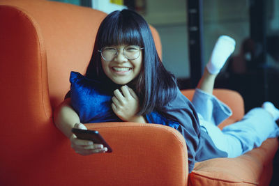 Asian teenager holding smartphone lying on sofa at home living room