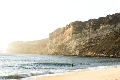 Scenic view of rocks and sea against clear sky with person surfboarding