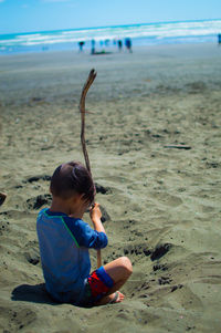 Boy with stick sitting at beach on sunny day