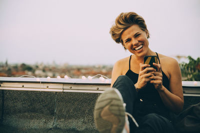 Portrait of cheerful woman using mobile phone while sitting on terrace against sky