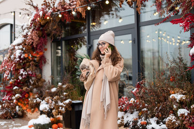 A smiling girl in a hat answers a call near a shop window in winter, holds a fir nobilis in a bag