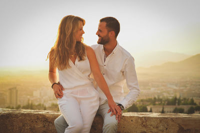 Young romantic couple sitting on retaining wall against sky during sunset