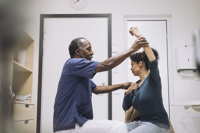 Side view of male doctor giving physical therapy to female patient sitting with hand raised in hospital