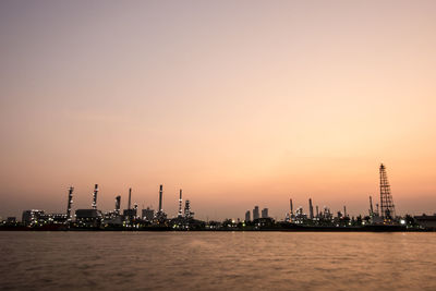 Illuminated factory by sea against sky during sunset