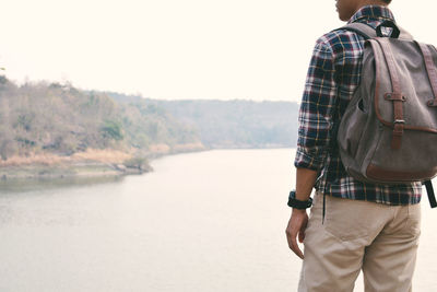 Midsection of man carrying backpack standing by lake against clear sky