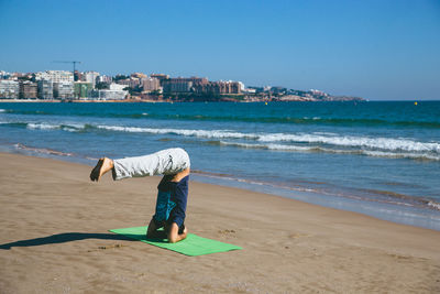 Full length of man performing handstand while doing yoga at beach against clear blue sky