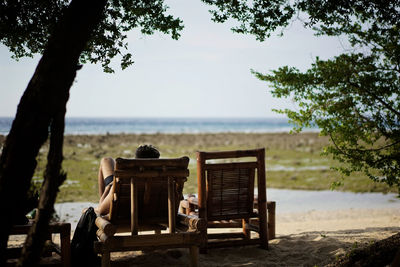 Rear view of man sitting on lounge chair at beach against sky