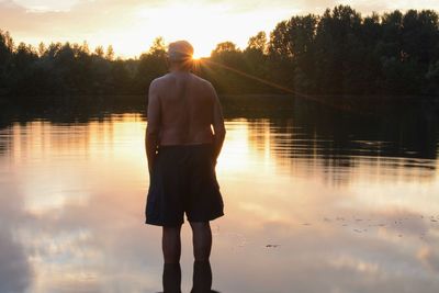 Rear view of man standing by lake against sky during sunset