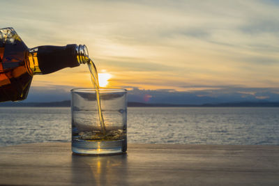 Bottle pouring whiskey in glass on table against sea during sunset