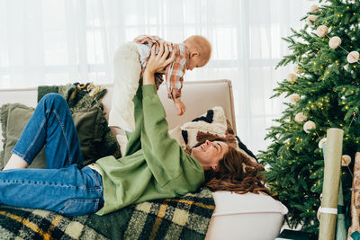 Young modern happy mom lying on the couch plays with the toddler, embraising and cuddling