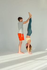 A slender woman stands on her hands, upside down, with the support of a teenager standing nearby.