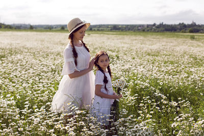 Mother with daughter in a white dress and hat stand in a daisy field in summer