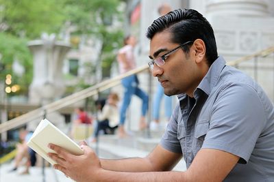 Young man reading book while sitting on steps in city