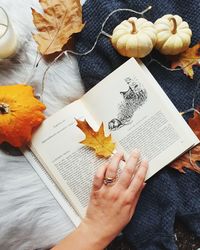 Cropped hand of woman holding book by pumpkins and dry leaves on bed